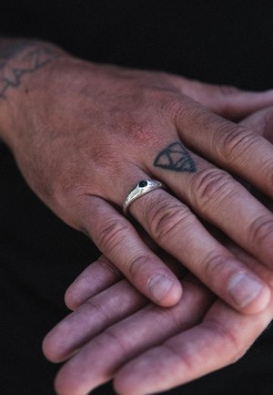 Silver ring with a dark gemstone, worn on a hand with tattoos, showing textured skin and pronounced knuckles. Hands are resting together.