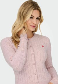 Svijetlo ružičasti pleteni kardigan s uzorkom pletenice, okruglim izrezom, šest tipki i malim crvenim srce detaljem na lijevoj prsnoj strani.