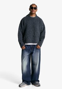 Sivi, teksturirani pleteni pulover; labave, baggy plave traperice s laganim izbljeđivanjem; sivi tenisice; crne sunčane naočale. Majica jednobojne boje ispod.