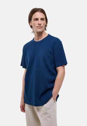 Homme aux cheveux bruns mi-longs portant un pull en maille bleu à manches courtes et un pantalon beige clair, debout avec une main dans la poche.