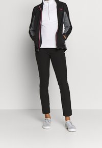 Svart idrottsjacka med grå detaljer och rosa dragkedja, kombinerad med en vit långärmad skjorta och svarta smala byxor. Ljusgrå skor.