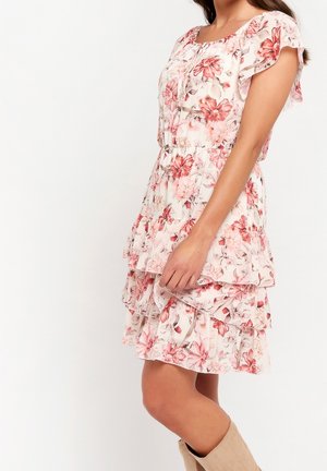 Femme portant une robe courte sans manches à fleurs avec des volants superposés, debout avec un bras détendu le long du corps sur un fond uni.