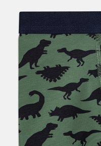 Kotystrikkede leggings med en mørkegrønn bakgrunn og svarte dinosaur-silhuetter i forskjellige poseringer og størrelser, samt et marineblått elastisk livbelte.