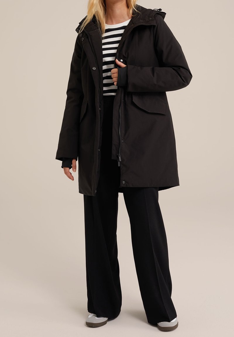 Czarna wodoodporna parka z zamkiem błyskawicznym i kapturem, wyposażona w dwie przednie kieszenie i luźny krój, noszona na paskowanym topie i szerokich spodniach.