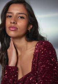 Burgunderfarbener, mit Pailletten besetzter Top mit tiefem V-Ausschnitt, strukturiertem Stoff und langen Ärmeln. Das Model hat dunkles Haar und berührt sanft ihren Hals.