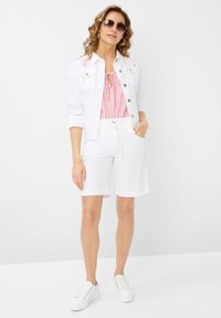 Veste en denim blanc sur un haut à rayures roses, accompagnée de shorts blancs et de baskets blanches. Le mannequin porte des lunettes de soleil et a les cheveux ondulés.