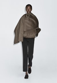 Model wearing a brown oversized wrap jacket, black tailored pants, and dark loafers walking against a plain light background.