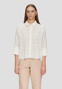 White button-up shirt with short sleeves, featuring eyelet embroidery, relaxed fit, and a collar. Pants are beige, made of a lightweight fabric.