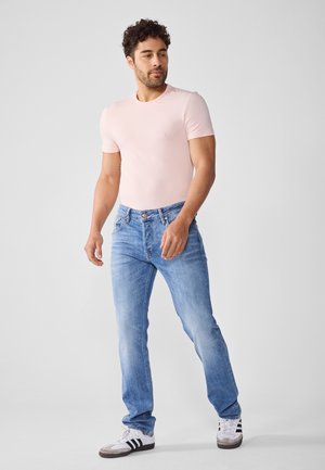 T-shirt de manga curta rosa claro, combinado com jeans azul desbotado e tênis brancos com detalhes em preto. O modelo está numa pose descontraída.