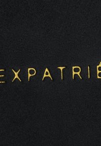Tessuto nero con testo in rilievo dorato che recita "EXPATRIÉ", mettendo in risalto una superficie liscia e un design elegante.