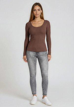 Kvinde står iført brun langærmet tætsiddende top, grå acid-vaskede skinny jeans og hvide sneakers mod ensfarvet baggrund.