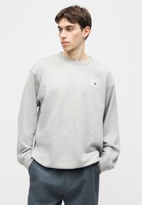 Νέος άνδρας με κοντά καστανά μαλλιά, που φορά ανοιχτό γκρι μακρυμάνικο φούτερ και σκουρόχρωμο παντελόνι, κοιτάζει προς τα αριστερά με τα χέρια στις τσέπες.