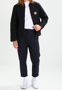 Musta vetoketjullinen takki, jossa etutasku, yhdistetty tummansinisiin housuihin. Valkoinen t-paita alla ja valkoiset lenkkitossut. Minimalistinen muotoilu ja istuvuus.