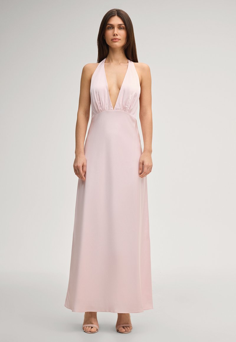 Frau steht und trägt ein ärmelloses, blassrosa Neckholder-Kleid mit tiefem V-Ausschnitt und bodenlangem Rock vor einfachem Hintergrund.