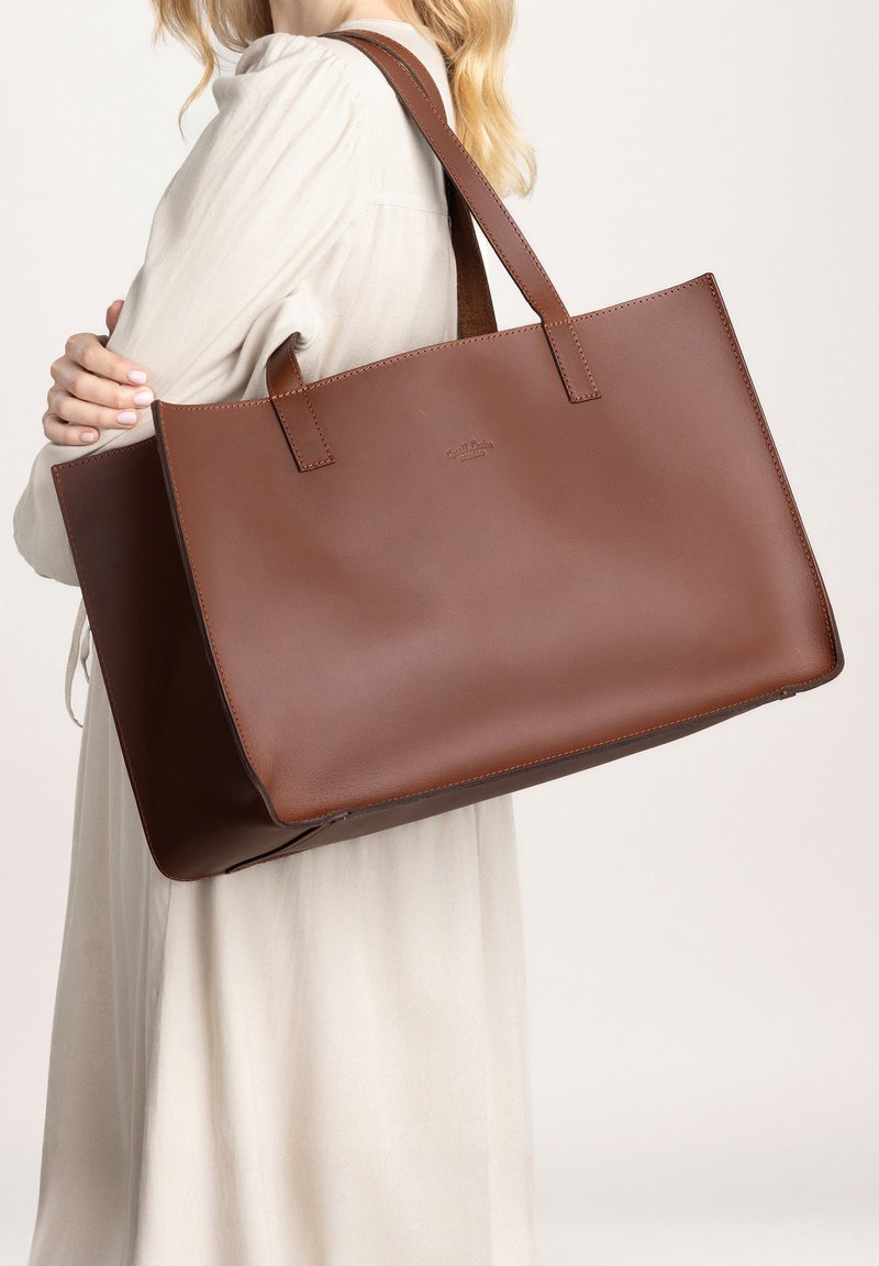 Sac fourre-tout en cuir brun avec une forme rectangulaire, des anses plates et un design minimaliste. Texture lisse avec un discret logo sur le devant.