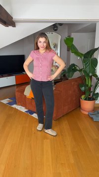 Młoda kobieta w czerwonej, kraciastej koszuli i czarnych spodniach stoi z rękami na biodrach w jasnym salonie z brązową kanapą i dużą zieloną rośliną.