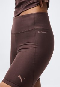 Κοντινό πλάνο ατόμου που φορά καφέ ποδηλατικές σορτς ψηλής μέσης με λογότυπο Puma και ετικέτα "Cloudspun" στο ισχίο.