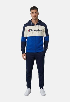Uomo che indossa una felpa con cappuccio Champion a blocchi di colore blu navy, bianco e blu, pantaloni della tuta navy e sneakers Champion bianche, in piedi con la mano in tasca.