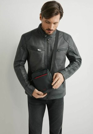 Mężczyzna w czarnej skórzanej kurtce trzyma małą czarną torebkę na ramię z czerwonym zamkiem na szarym tle.