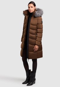 Rjava puhasta bunda z umetnim krznenim kapuco, ki ima oprijet kroj, horizontalno prešivanje in žepke z zadrgo. Nosena s črnimi gležnjarji.