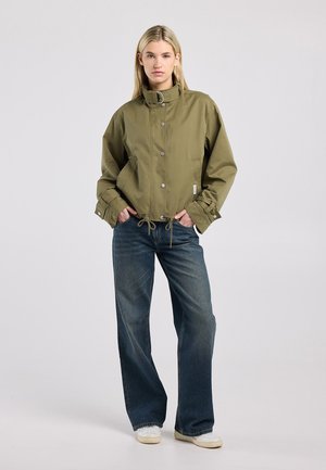 Vrouw staand met handen in de zakken, draagt een olijfgroene jas met knopen, blauwe jeans met wijde pijpen en witte sneakers tegen een effen achtergrond.