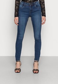 Mulher vestindo jeans skinny azul escuro, blusa de renda preta de mangas longas e sandálias de salto altas pretas com tiras, apoiada contra um fundo simples.
