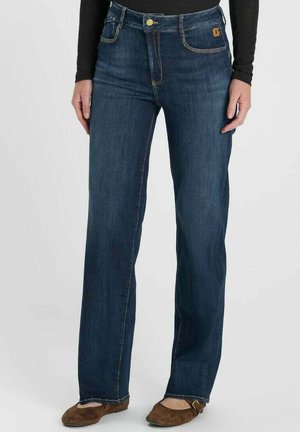 Donna che indossa jeans blu scuro a gamba dritta, top nero a maniche lunghe e scarpe basse marroni, in piedi davanti a uno sfondo semplice.
