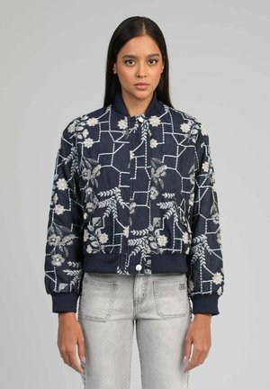 Femme portant une veste bomber bleu marine avec une broderie florale blanche et un jean denim gris clair, debout devant un fond gris uni.