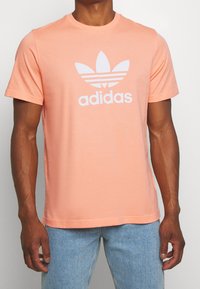 Ljusrosa bomullst-shirt med rund hals och korta ärmar, prydd med en stor vit Adidas-logotyp och treklöverdesign på framsidan.