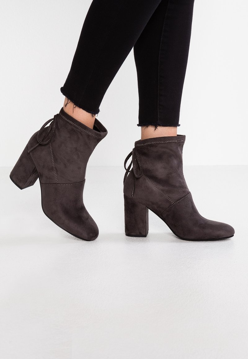 Bottes en daim marron foncé montant à la cheville avec talons carrés et nœuds à l'arrière, portées avec un pantalon skinny noir à ourlets effilochés sur un fond blanc.