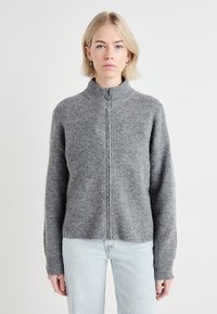 Pull en zip gris avec un col haut et une texture côtel�ée. Présente des manches longues et une coupe décontractée. Porté avec un jean bleu clair.