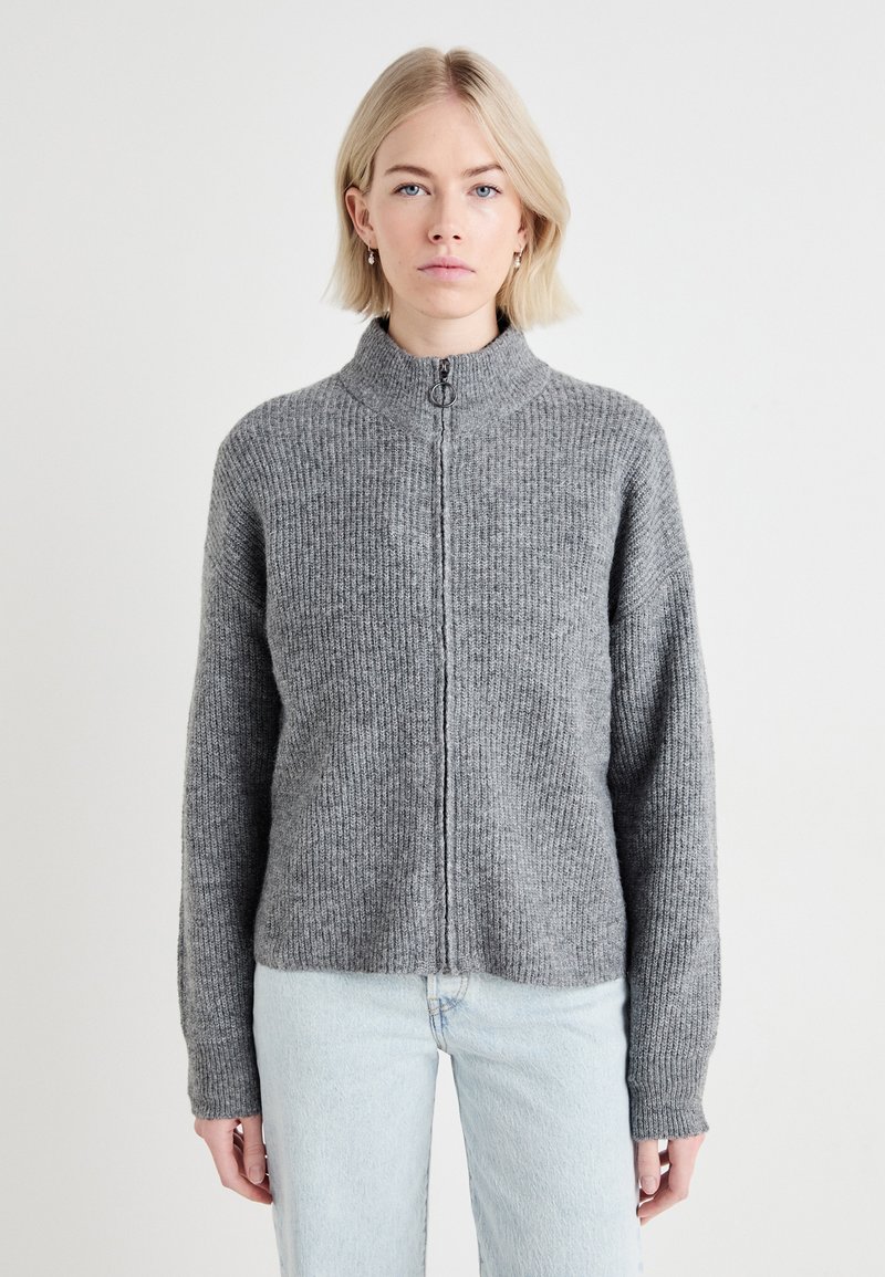 Pull en zip gris avec un col haut et une texture côtelée. Présente des manches longues et une coupe décontractée. Porté avec un jean bleu clair.