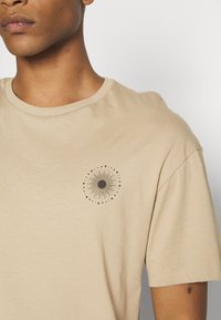 Beige Baumwoll-T-Shirt mit Rundhalsausschnitt, das ein zentrales schwarzes kreisförmiges grafisches Design mit strahlenden Linien und Punkten zeigt.
