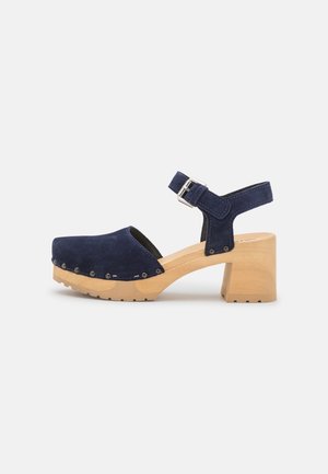 Marineblaue Wildleder-Clog-Sandale mit Holzblockabsatz, Knöchelriemen, Metallschnalle und Nietenverzierung entlang der Sohlenkante.