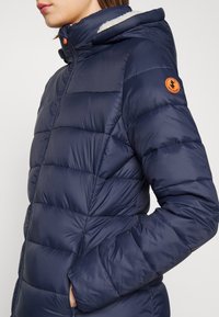 Merinopuffer takki tummansinisellä värillä, jossa on teksturoitu pinta, huppu, etuvetoketju ja oranssi logopatchi vasemmassa hihassa.