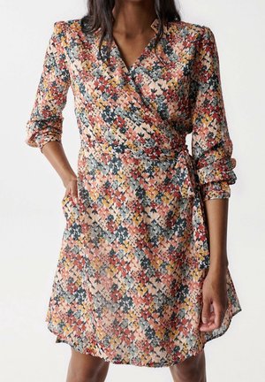 Femme portant une robe portefeuille fleurie mi-longue avec des manches trois quarts dans des nuances de rouge, jaune, gris et noir, une main dans la poche.