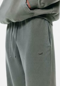 Grå bomulds sweatpants med elastisk talje og snor, med et lille sort logo på venstre lår. Glat tekstur, uformelt design.