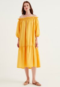 Gul off-the-shoulder klänning med rynkad halsringning, voluminösa ärmar och ett lager på lager nederdel. Mjuk tyg med slät textur. Bärs med platta skor.