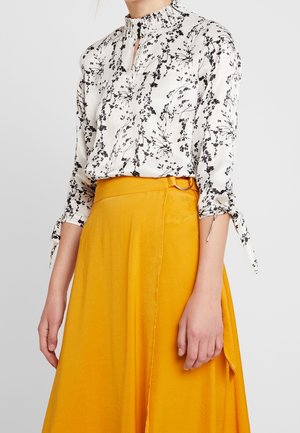 Mujer con blusa blanca de manga larga con estampado floral negro y falda amarilla de talle alto con cinturón de tela y aro metálico lateral.
