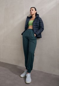 Schwarze Pufferjacke, eng anliegendes grünes Crop-Top, dunkelgrüne Jogginghose und hellgraue Sneakers mit neonfarbenen Akzenten. Lässiges Sportoutfit.