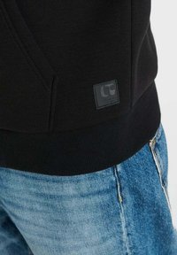 Zwarte hoodie met een geribde zoom en zijzakken. Voorzien van een getextureerde rechthoekige merkpatch met een geperst logo. Gecombineerd met blauwe denim.