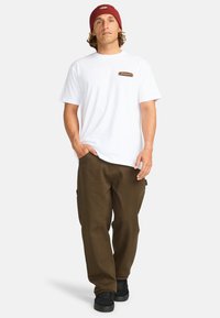 Hvid t-shirt med korte ærmer og et brunt logo-patch, kombineret med brune cargo-bukser og sorte sneakers. Personen bærer en rød beanie.