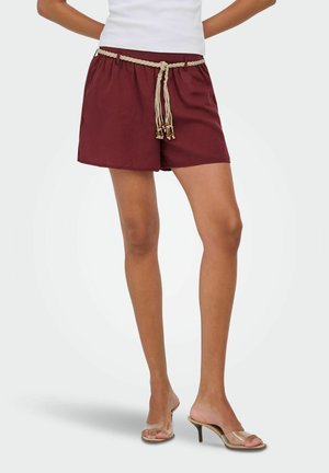 Femme portant un short bordeaux avec une ceinture tressée beige et des sandales à talons hauts avec brides transparentes, debout les mains sur les hanches.