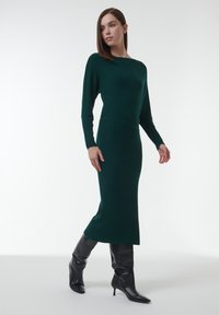 Femme portant une robe midi en tricot côtelé à manches longues de couleur vert foncé et des bottes à talons montantes noires, se tenant devant un fond blanc.