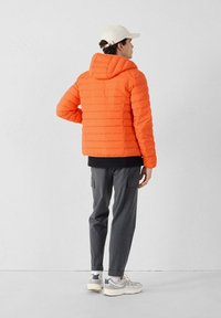 Chaqueta de plumas naranja brillante con costuras horizontales, capucha y puños acanalados. Lleva pantalones cargo grises y zapatillas grises, de espaldas al espectador.