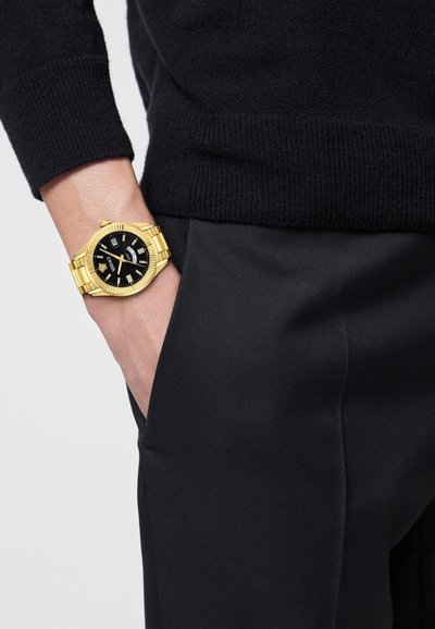Orologio in acciaio inox dorato con quadrante nero, dettagli in argento e display della data, indossato su un polso con un maglione scuro e pantaloni neri.
