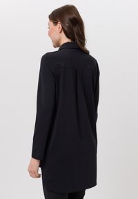 Schwarze Tunika mit Kragen, langen Ärmeln und einer Rückenfalte. Hergestellt aus glattem Stoff mit leicht geschwungenem Saum.