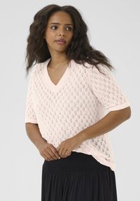 Light pink knitted sweater featuring a V-neck, short sleeves, and open diamond pattern. Paired with a black smocked skirt.