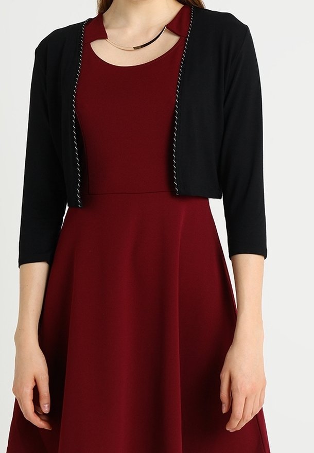 Femme portant une robe sans manches bordeaux avec un décolleté découpé et un cardigan noir court à manches trois quarts.