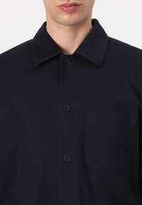 Homme portant une chemise bleu marine foncé à boutons, avec un col et une poche poitrine, montré du cou jusqu'au milieu du torse.